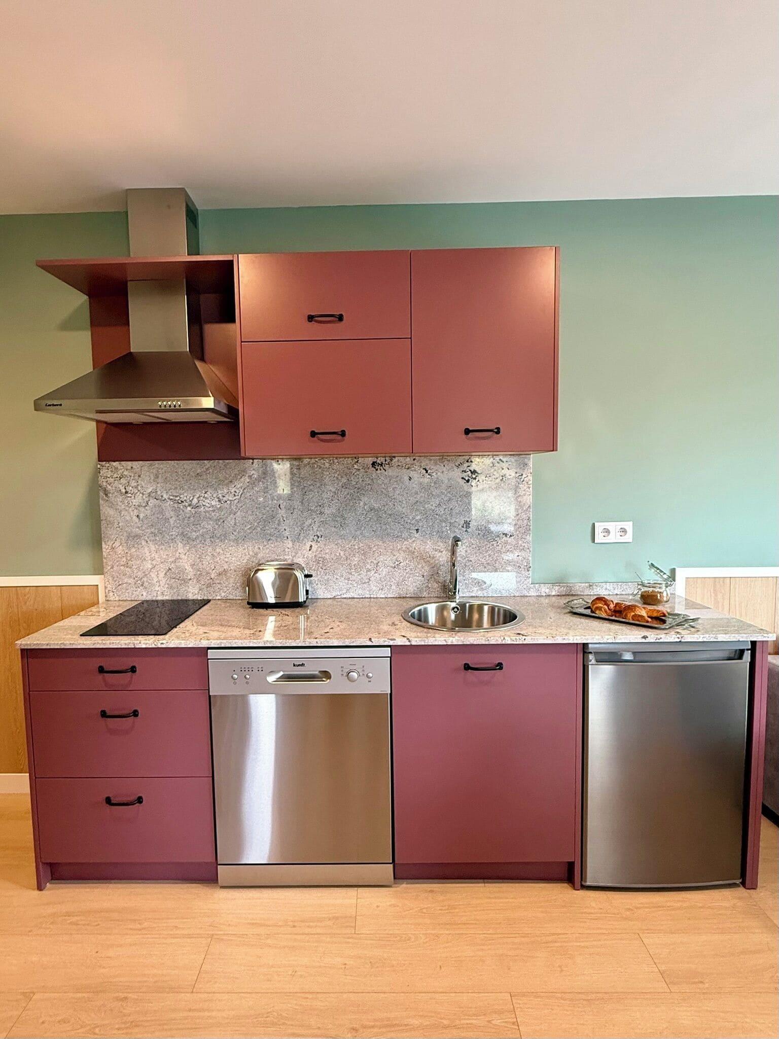 A modern kitchen with stainless steel appliances, a granite countertop, and a teal wall.