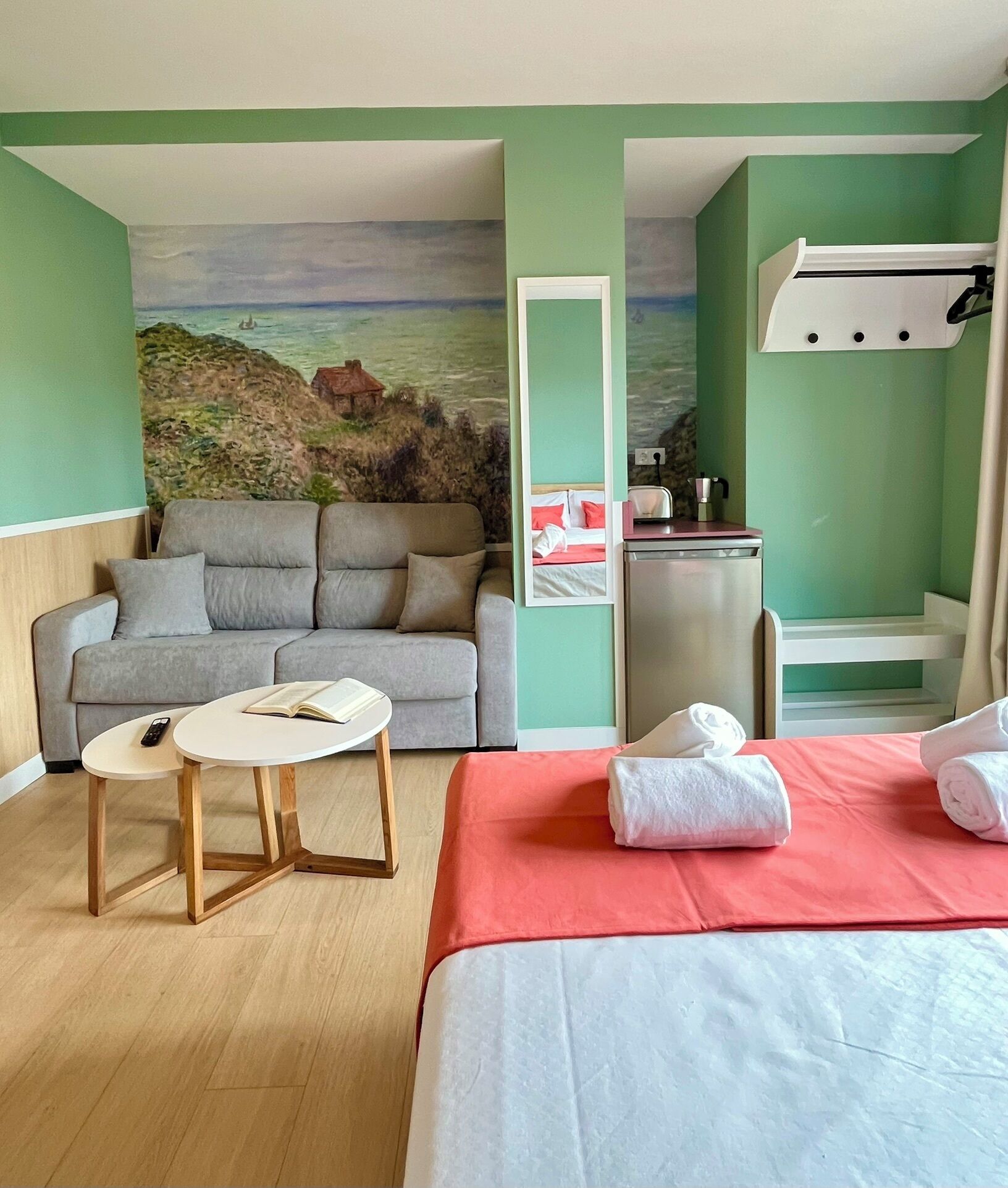 Habitación de hotel con cama, sofá, una mesita, espejo, escritorio y vista al mar a través de un mural.