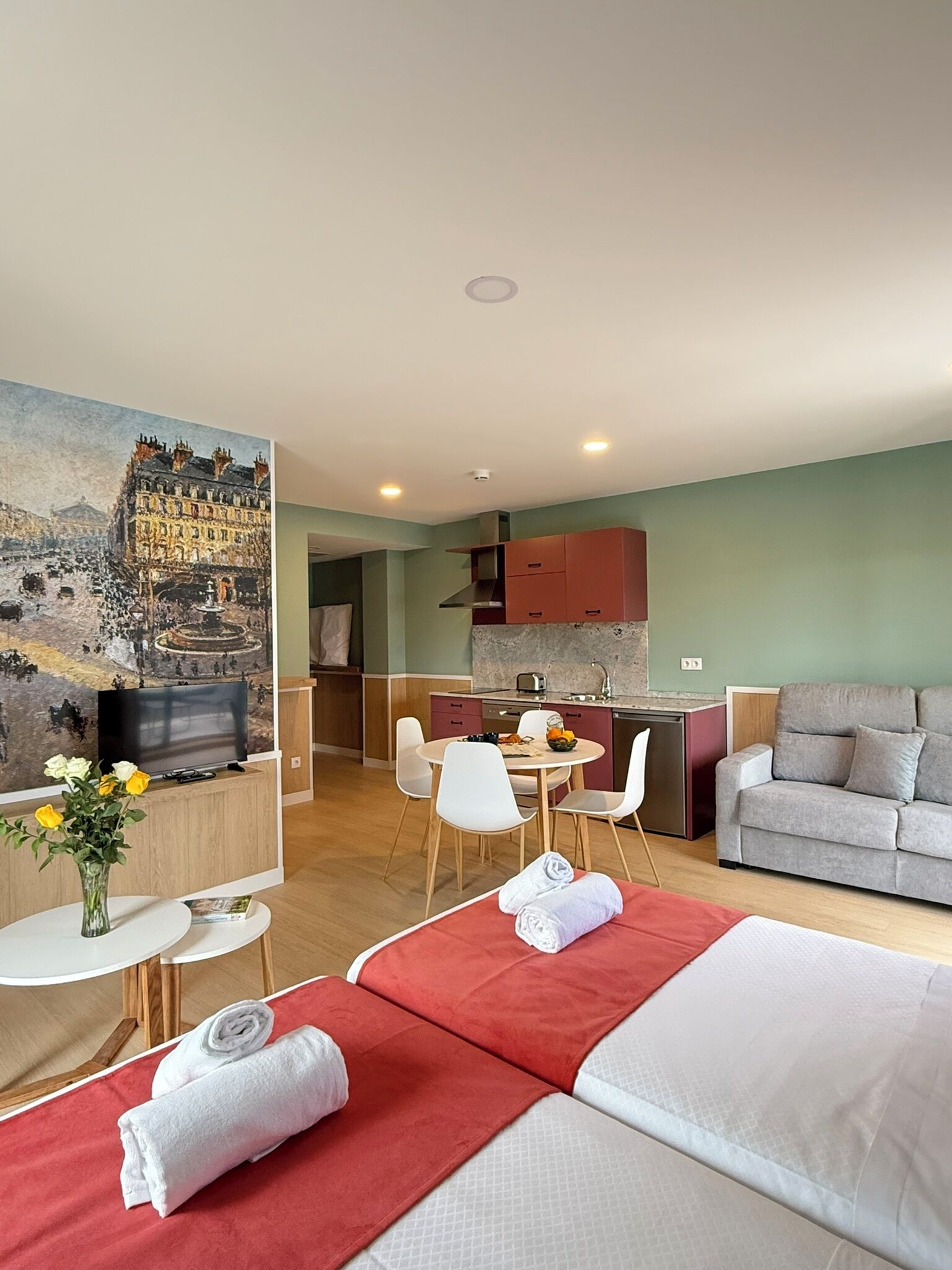 A hotel room with a bed, a TV, a dining table with chairs, a kitchen area, and a painting on the wall.