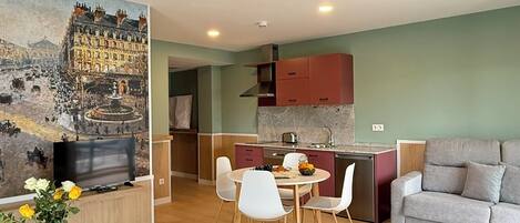 A hotel room with a bed, a TV, a dining table with chairs, a kitchen area, and a painting on the wall.