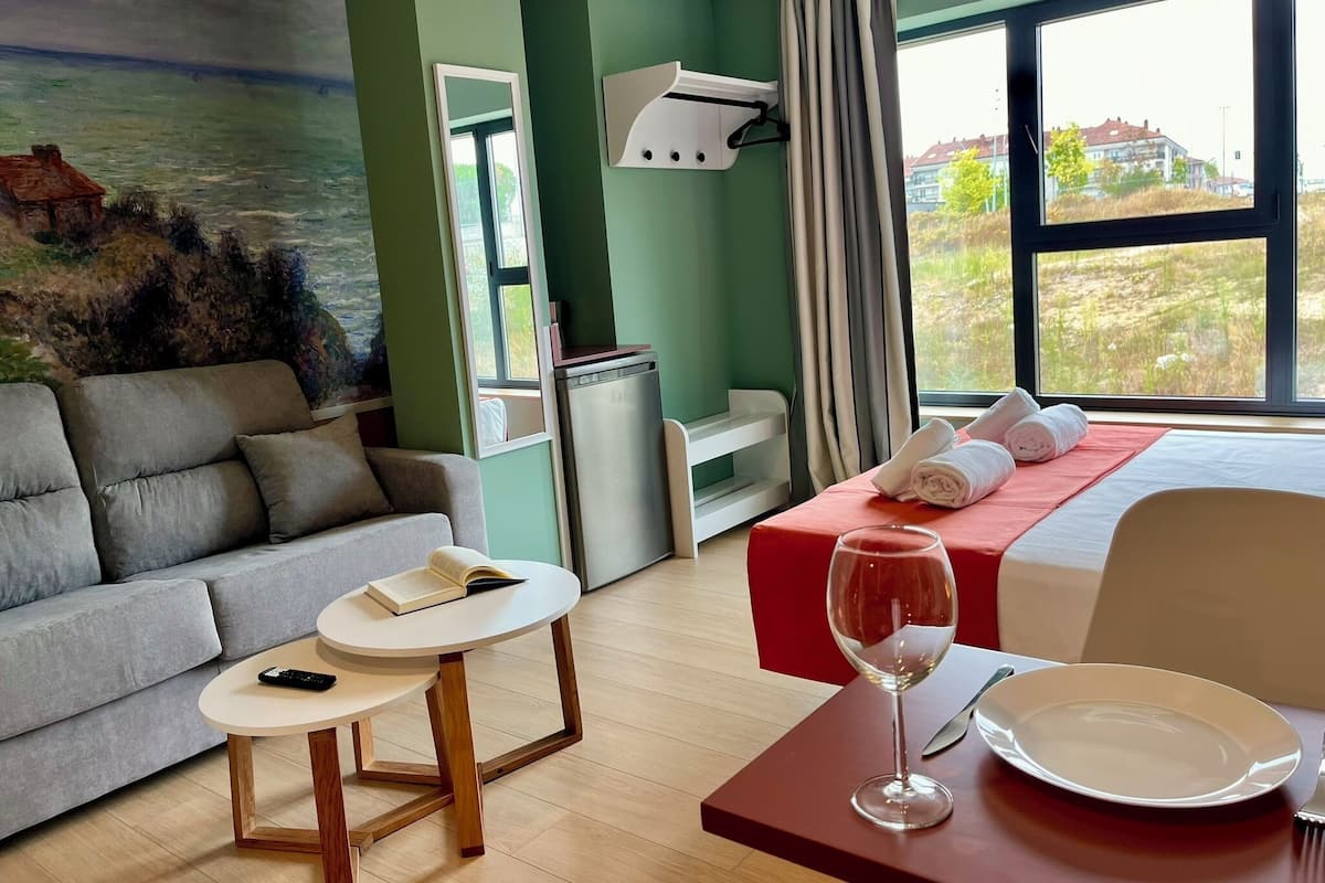 A hotel room with a bed, a sofa, a small table, a desk, a window with a view, and a painting on the wall.