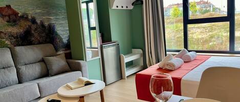 Habitación de hotel con cama, sofá, una mesita, escritorio, ventana con vistas y un cuadro en la pared.
