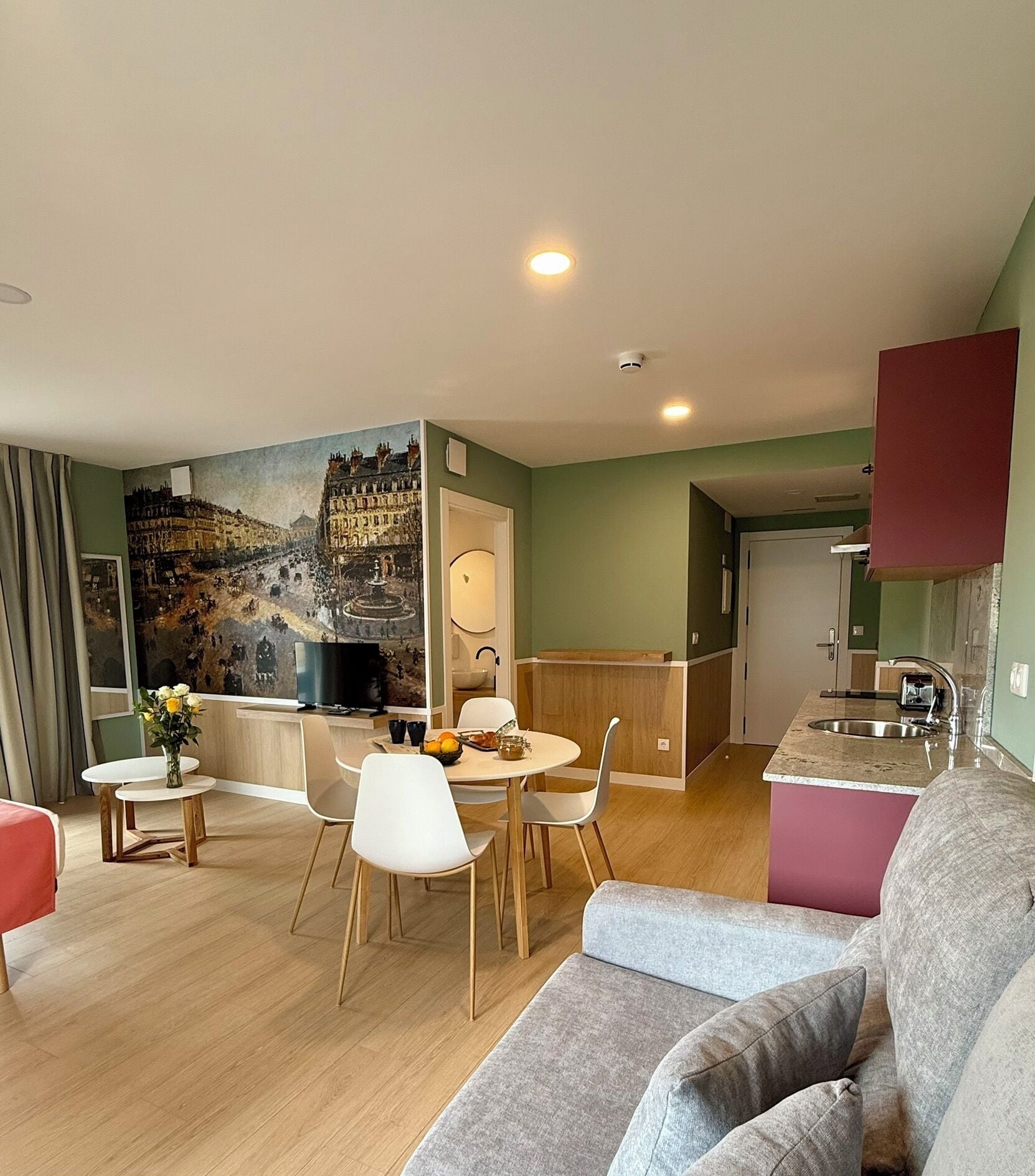 Una sala de estar moderna con un sofá gris, una zona de comedor con una mesa y sillas blancas, una cocina con un fregadero y alacenas, y un mural en la pared.