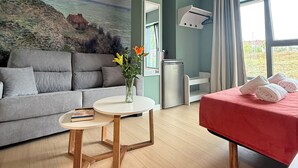 Une chambre d’hôtel moderne avec un canapé gris, une petite table ronde, un lit rouge et un tableau représentant un paysage côtier accroché au mur.