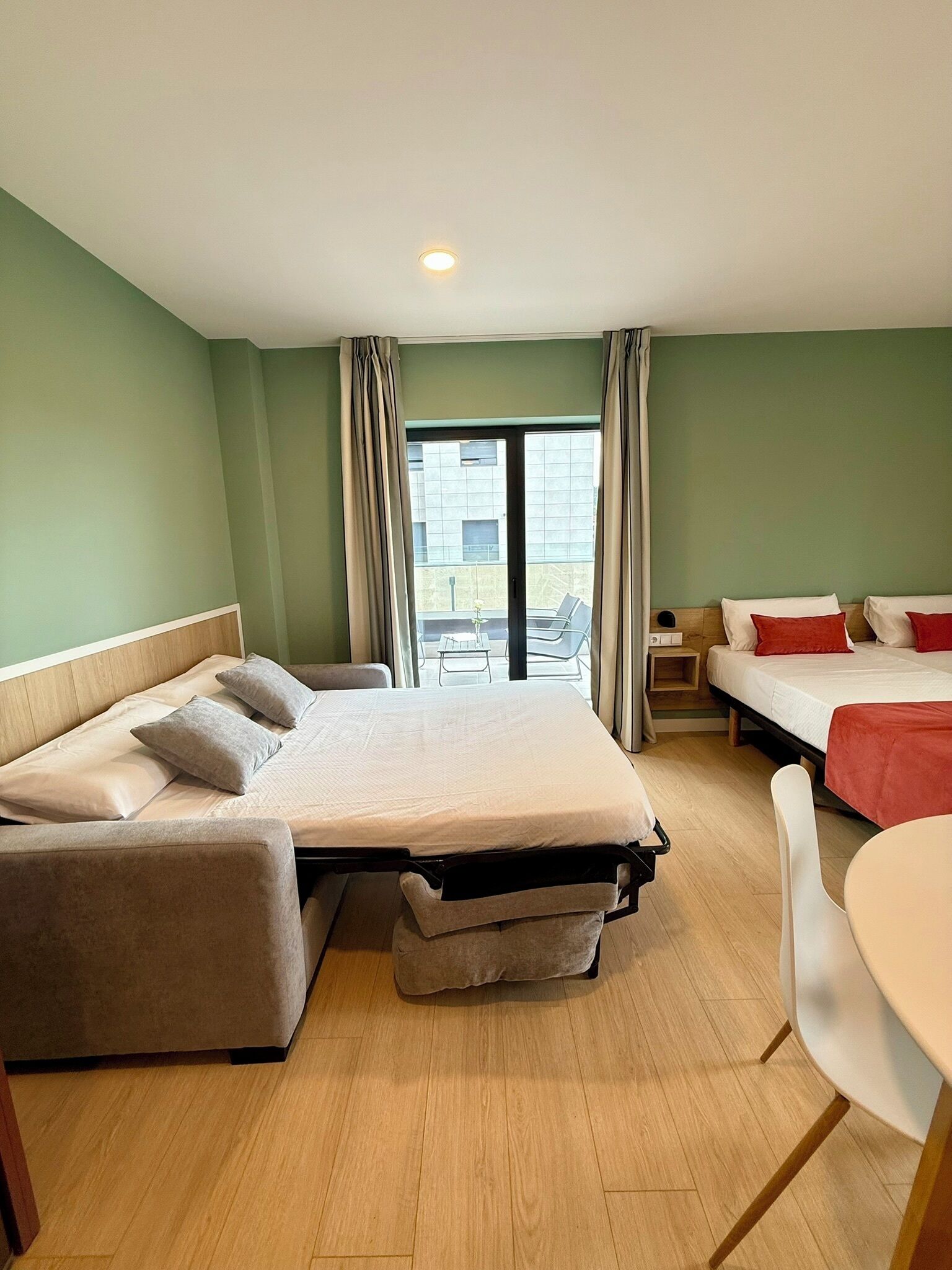 A hotel room with a bed, a sofa, a desk, and a chair. There is a window with curtains and a view of the outside.