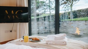A neatly made bed with a glass of orange juice, a croissant, and folded towels, a stone wall visible through a glass door, and a fireplace in the background. - New Peaceful Countryside Annex (Leyburn)
