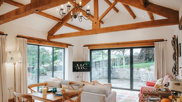 A spacious living room with a high ceiling, wooden beams, a large TV, and a dining area with a table and chairs. - New Peaceful Countryside Annex (Leyburn)