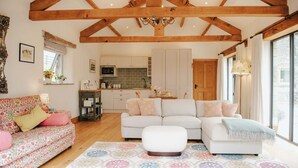 A cozy living room with a sofa, ottoman, and a patterned rug. A kitchen with white cabinets and a microwave is visible in the background. - New Peaceful Countryside Annex (Leyburn)