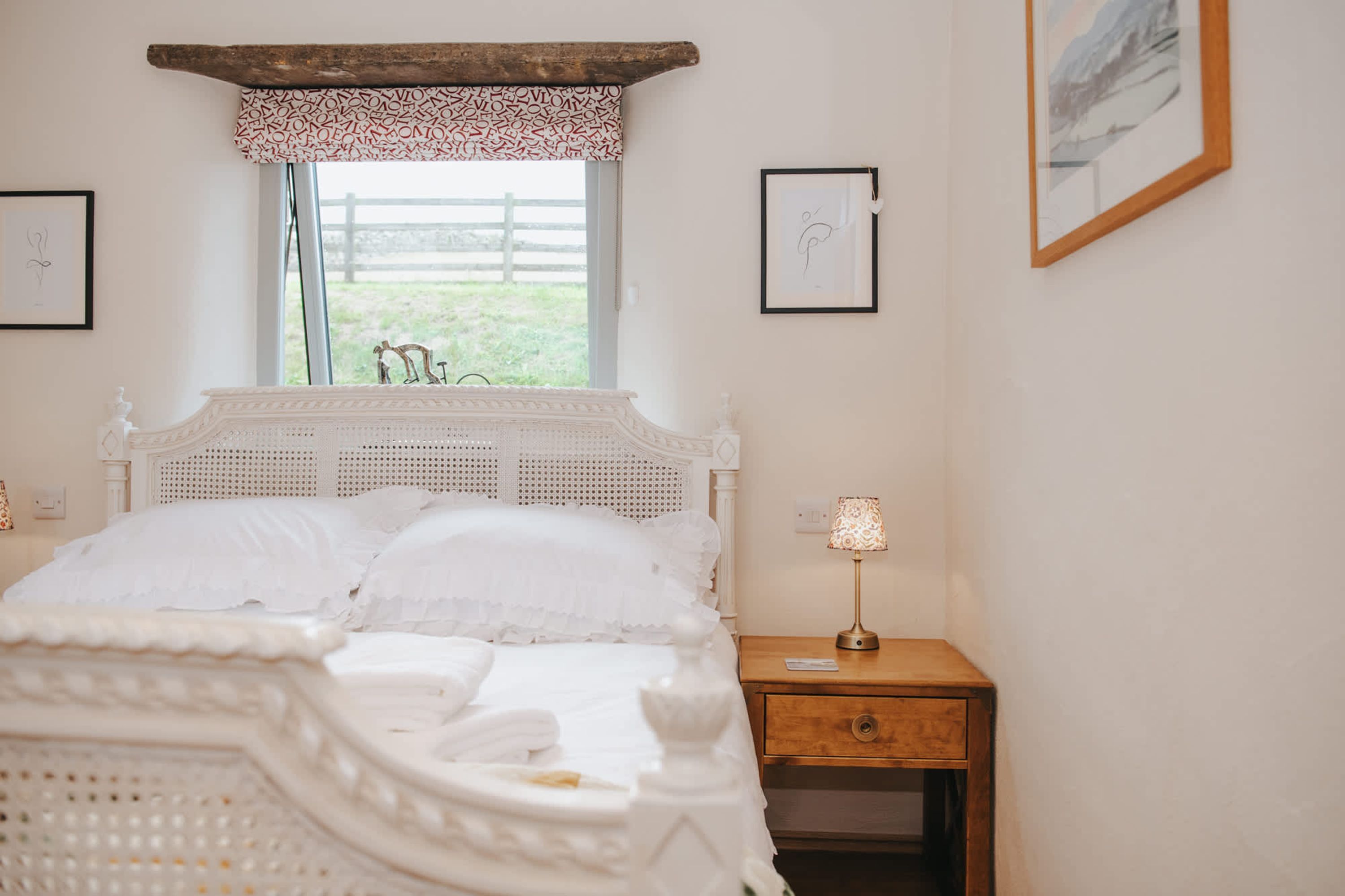 A bedroom with a white bed, a wooden nightstand, and a window with a view of a fence and greenery.