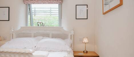 A bedroom with a white bed, a wooden nightstand, and a window with a view of a fence and greenery.