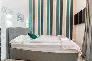 A bed with a grey headboard, white bedding, and a green pillow. A striped wall pattern is visible behind the bed.