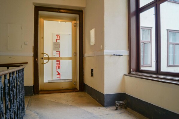 A hallway with a door marked 'FLAT TIME AT,' a window with a view of buildings, and a metal railing.