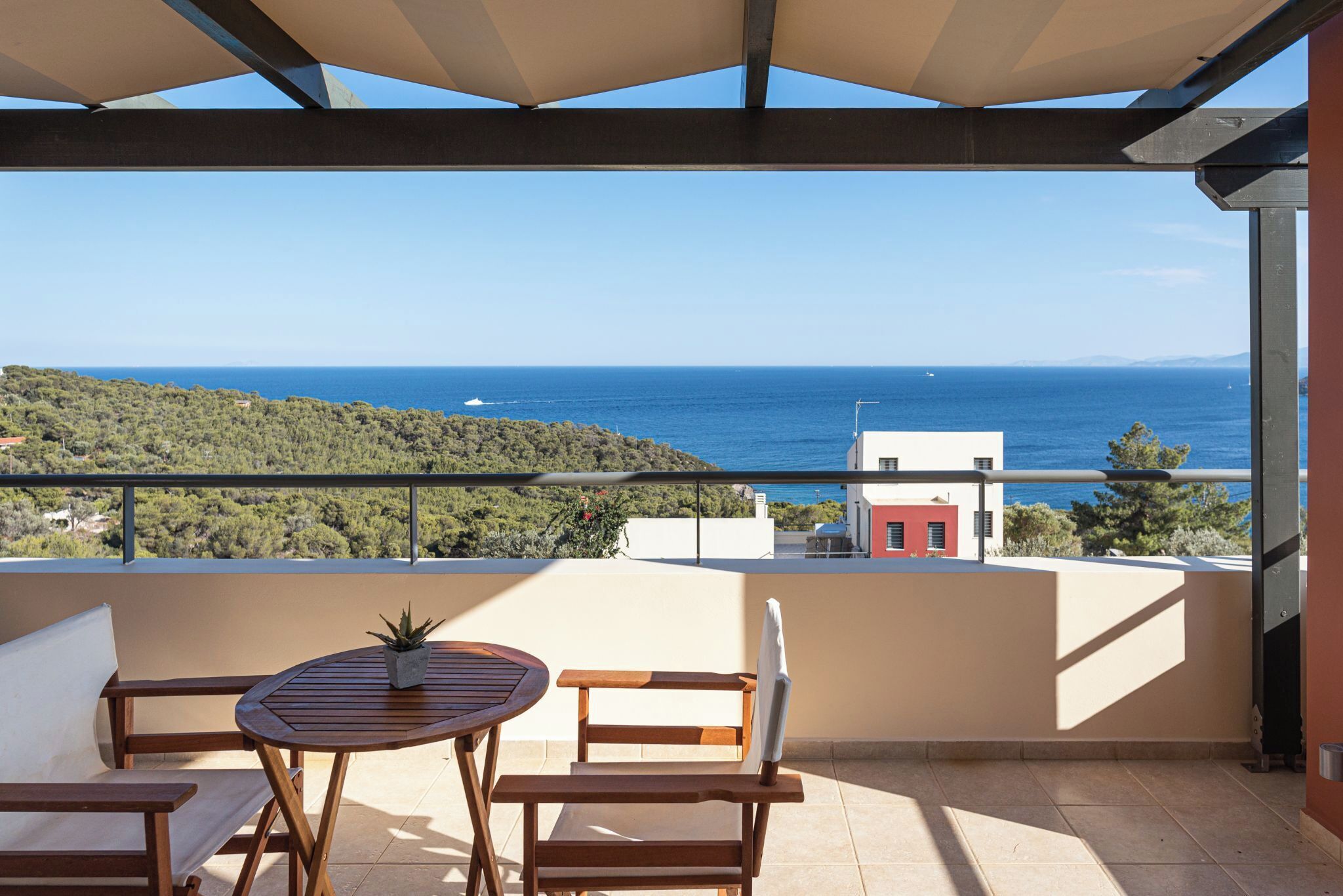 Un balcón con una mesa y sillas de madera, con vistas a la costa, el mar y colinas distantes.