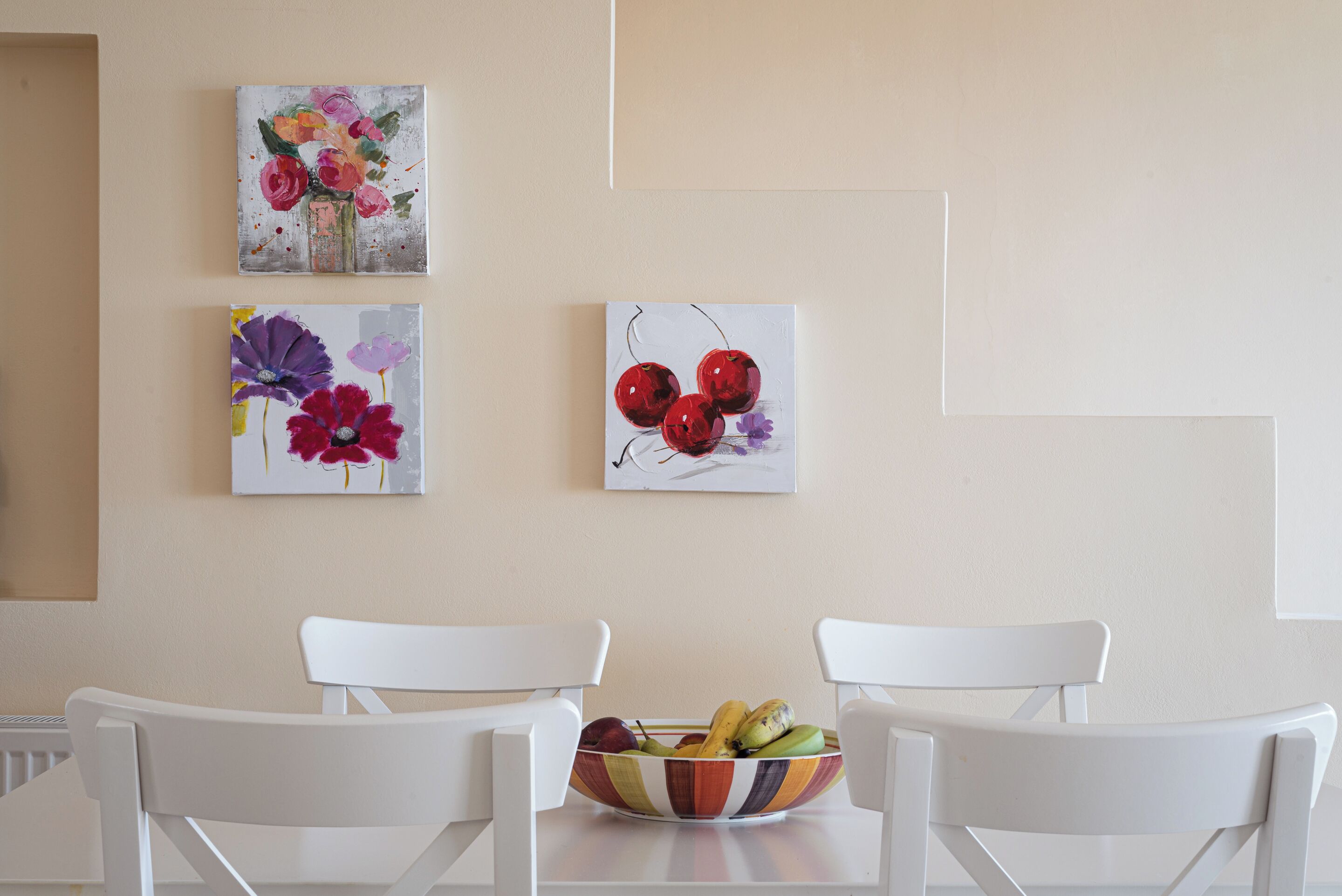 Ein Essbereich mit weißen Stühlen, einem Tisch und einer Obstschüssel. An der Wand hängen drei Gemälde.