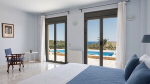 A bedroom with a large bed, a chair, a small table, and a view of an outdoor swimming pool. - Aphaias Residence Two, Aphaias Villas (Aegina)