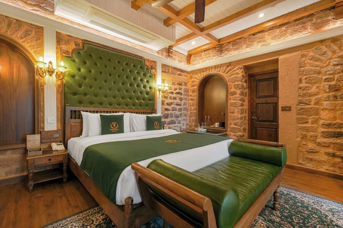 Un dormitorio con una cama grande, un cabecero tapizado de color verde, un banco de madera y paredes de piedra.