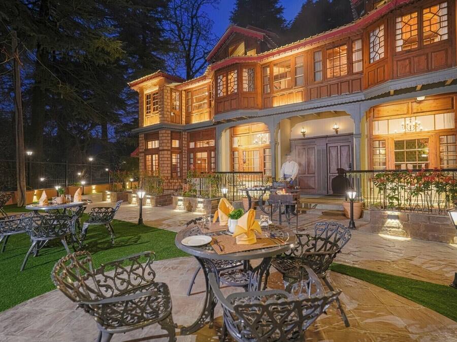 An outdoor dining area with tables and chairs, a chef preparing food, and a building with large windows and a balcony.