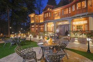 An outdoor dining area with tables and chairs, a chef preparing food, and a building with large windows and a balcony.