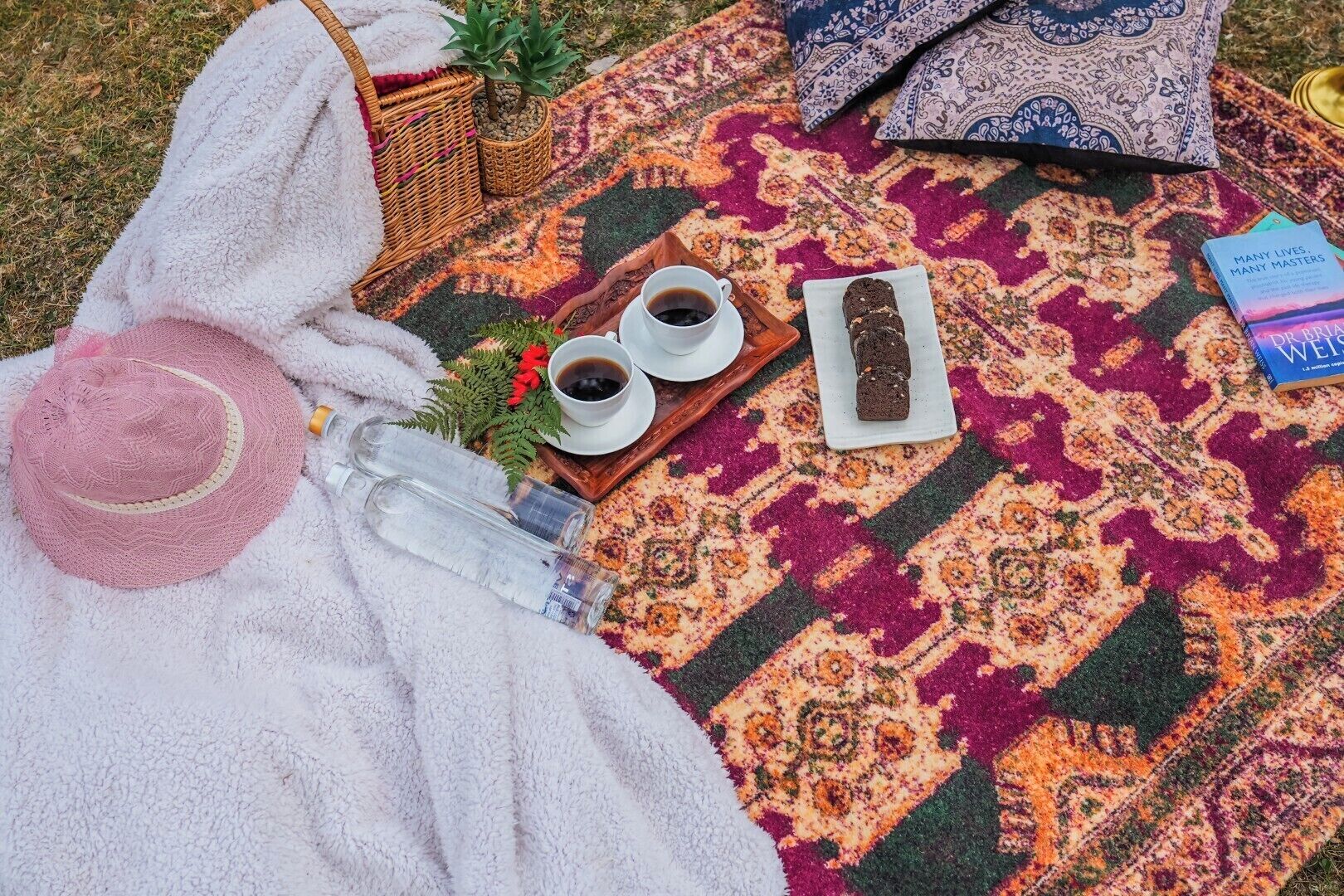 Un montaje para un picnic, con una manta, un sombrero, una bandeja con bebidas, un libro y una botella.