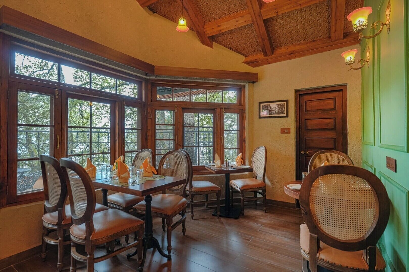 Un comedor con mesas y sillas de madera, ventanas amplias con vistas a árboles y una pared verde con una puerta.