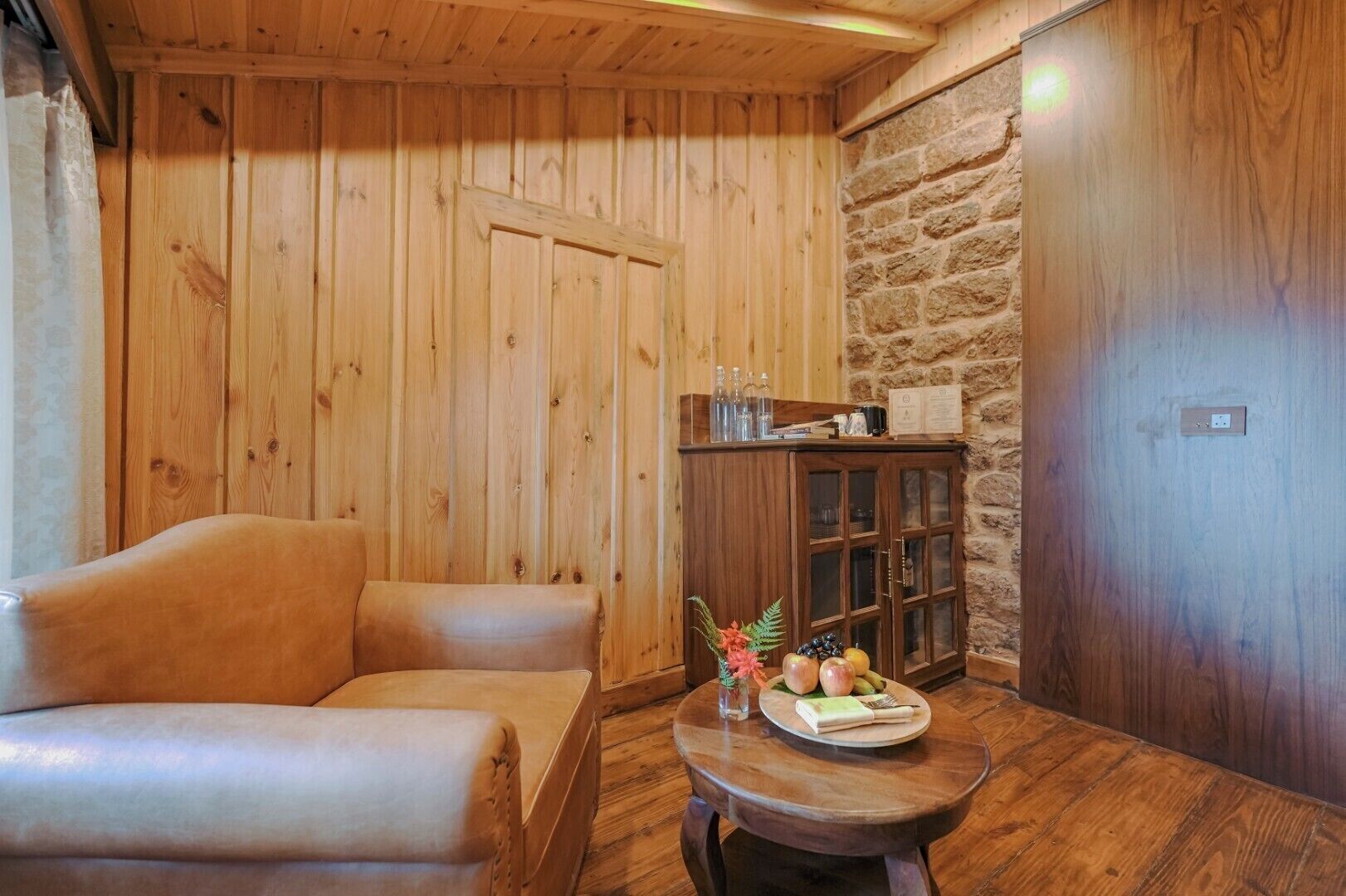 Habitació acollidora amb parets de fusta, una paret decorativa de pedra, un sofà amb coixins, una taula petita amb fruita i un llibre, i un armari amb portes de vidre.