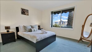 Une chambre à coucher comprenant un lit, une table de chevet, une commode, un miroir et une fenêtre donnant sur des maisons.