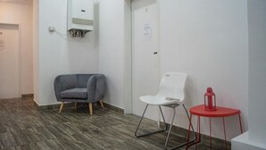 Um corredor com uma poltrona cinzenta, uma cadeira branca e uma mesa vermelha com um candeeiro.