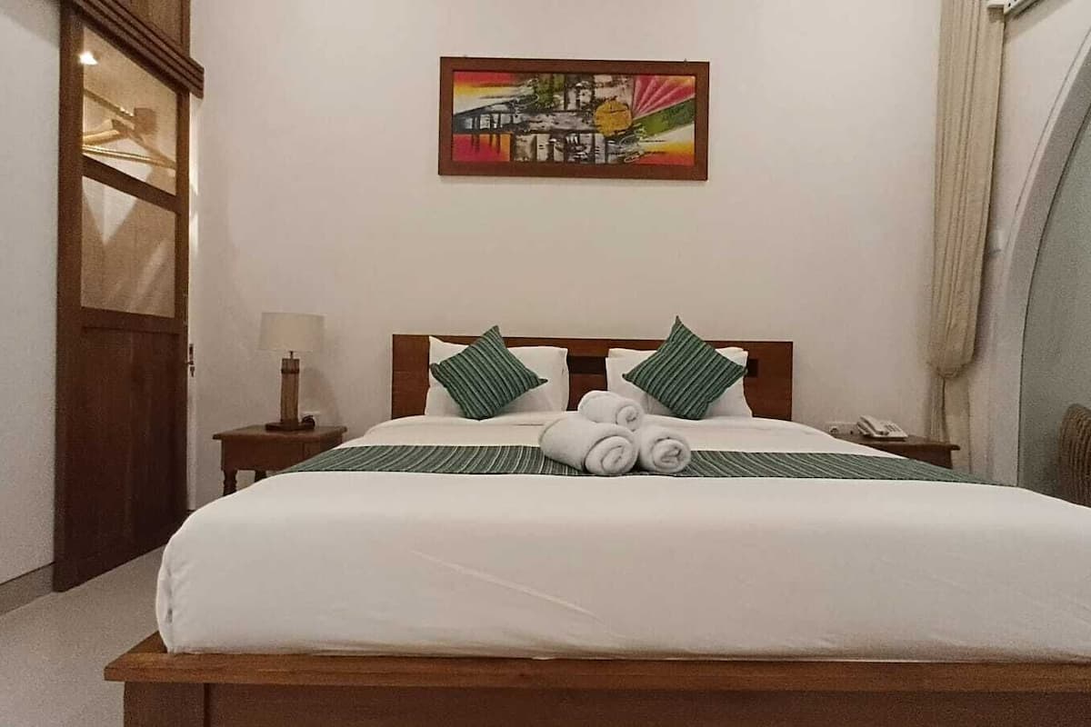 Una cama bien tendida con sábanas blancas y almohadas a rayas verdes, un cabecero de madera y un cuadro en la pared.