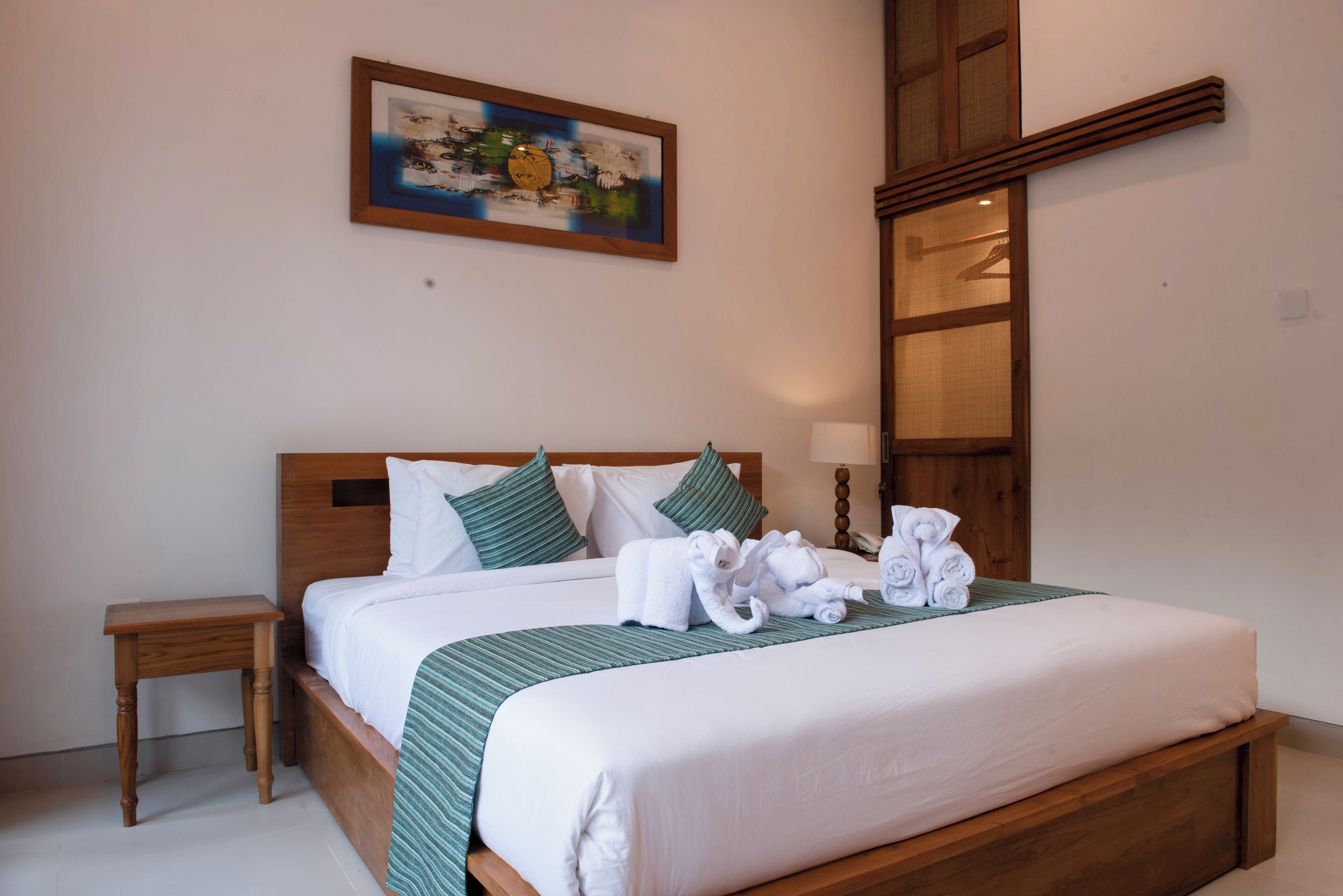 A neatly made bed with white linens and green accents, a small wooden nightstand, and a painting on the wall.