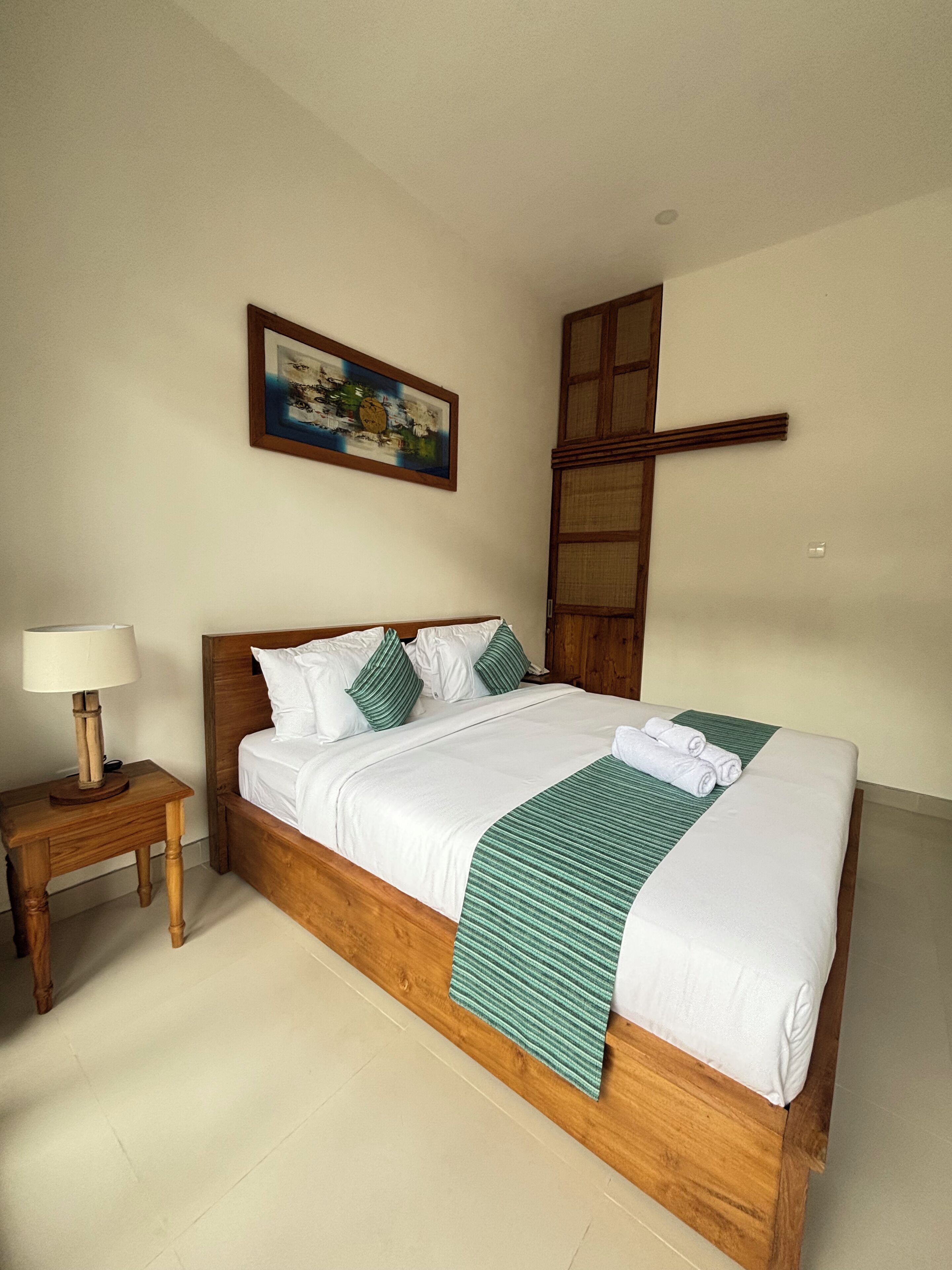 Una cama bien tendida con sábanas blancas y detalles en verde, una mesita de noche de madera con una lámpara, y un cuadro en la pared.