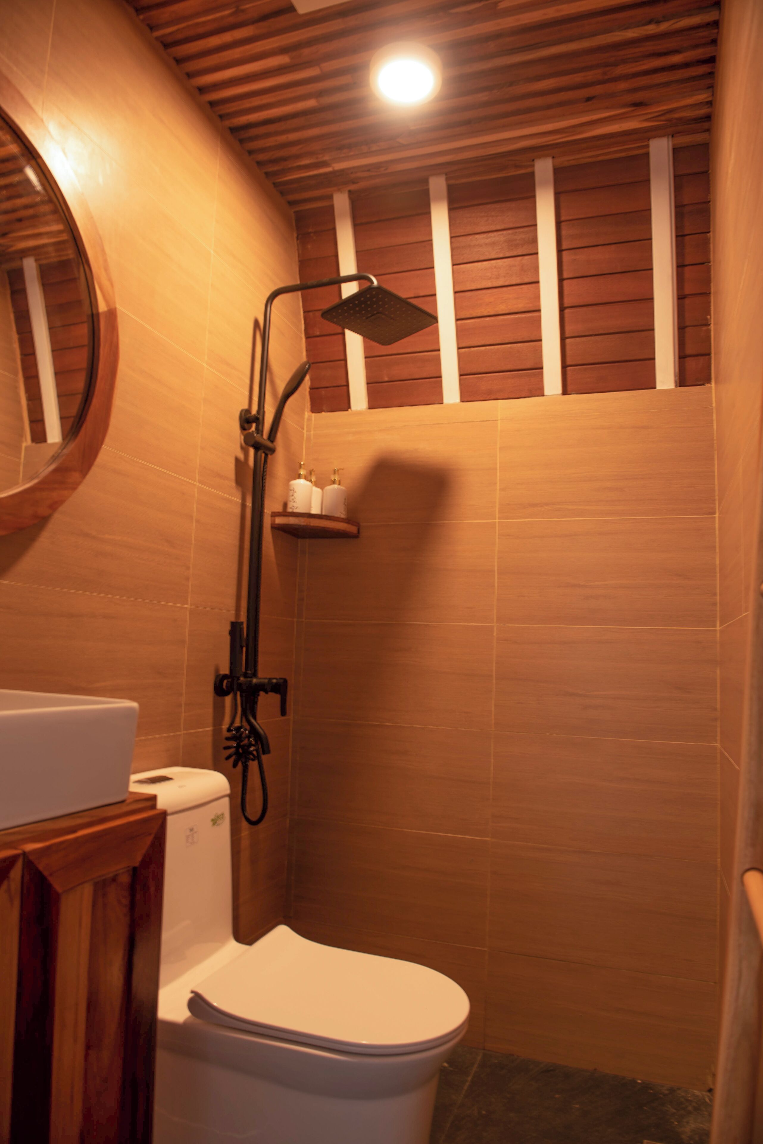 A bathroom with a wooden ceiling, a white toilet, a shower with a handheld showerhead, and a shelf with toiletries.