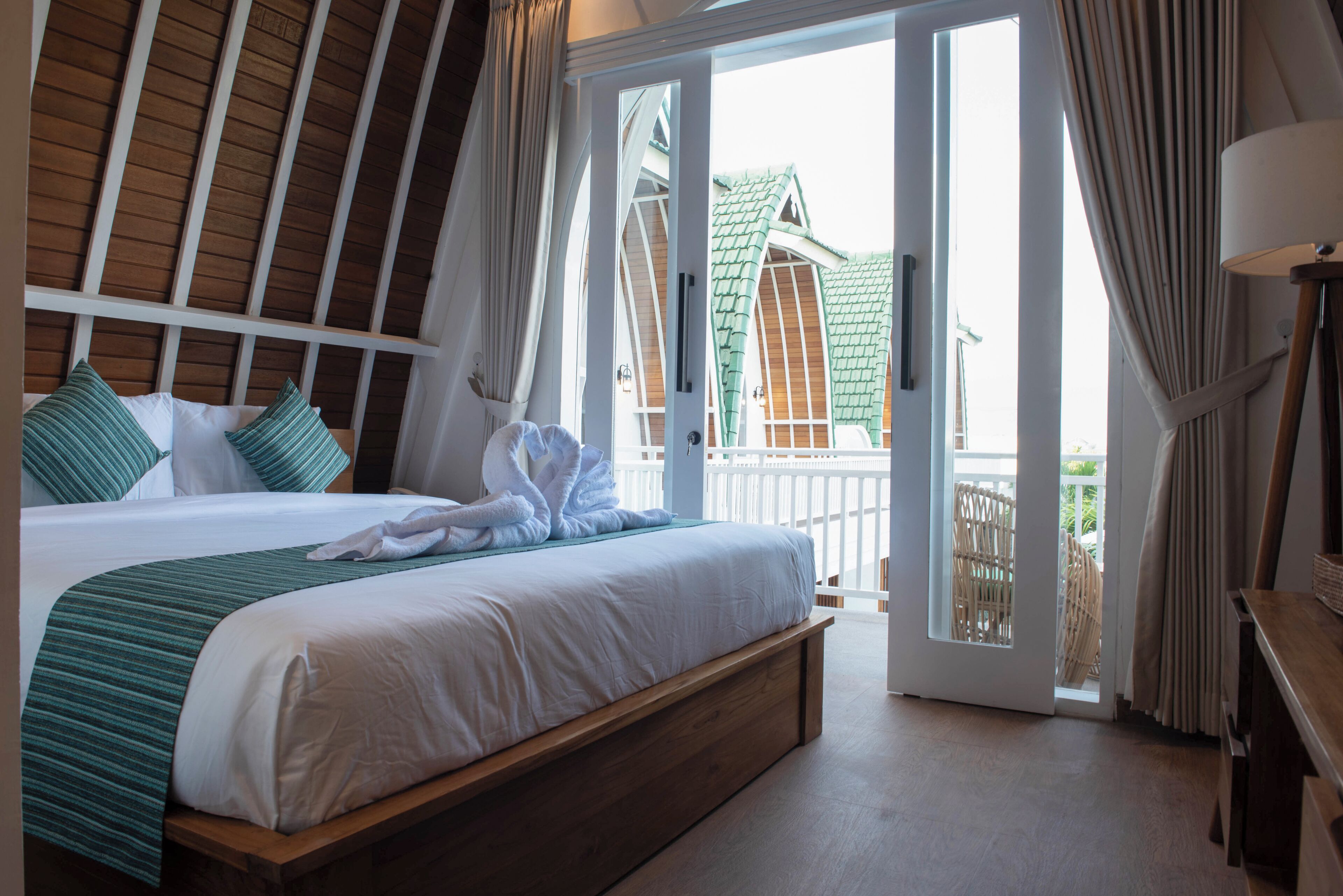 Habitación de hotel con una cama grande, cabecera de madera, balcón con vista a un edificio y una lámpara en una mesita de noche.