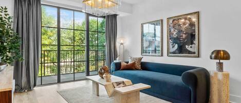 A modern living room with a blue sofa, a wooden coffee table, a hanging light fixture, and large windows with a view of greenery.