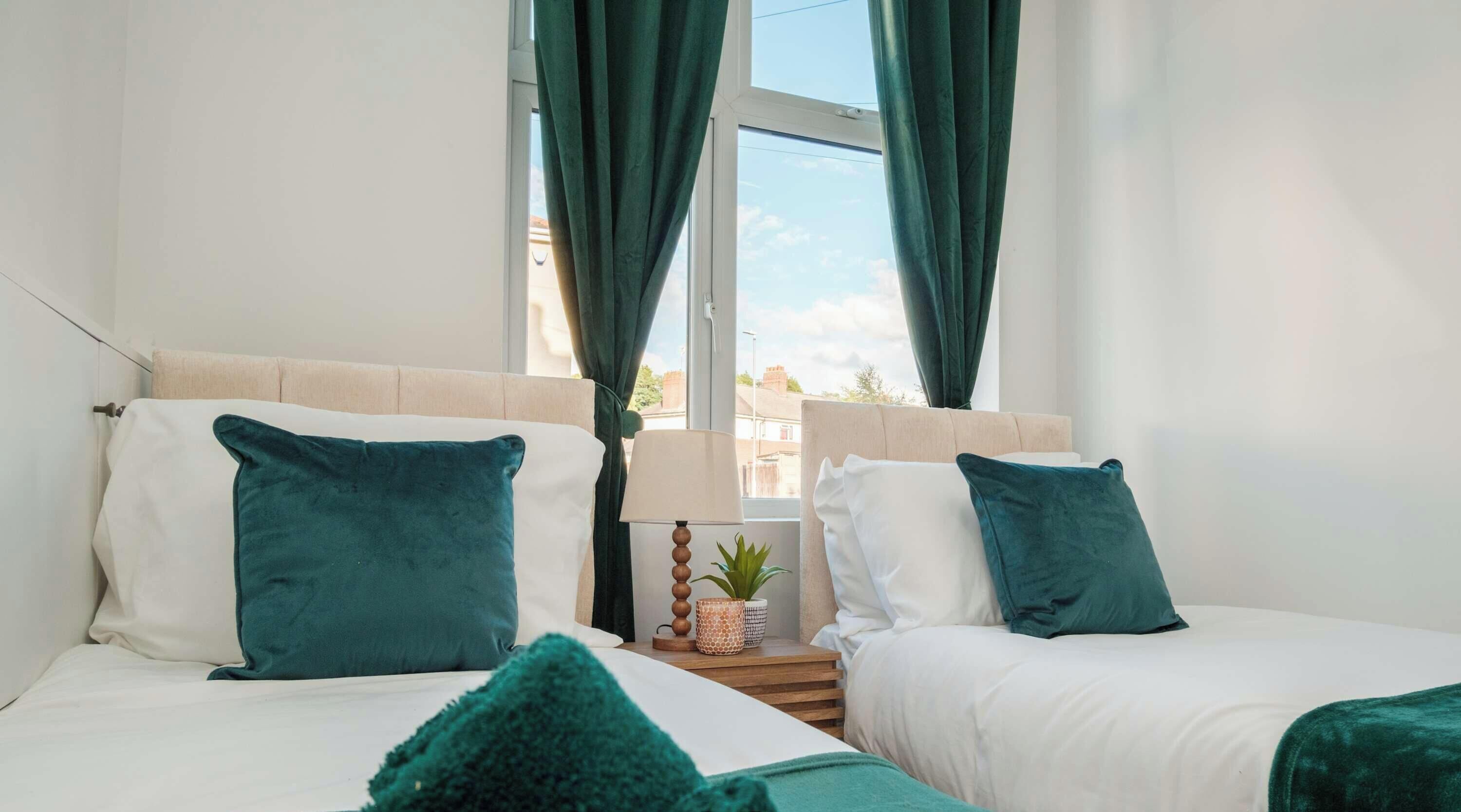 A hotel room with two beds, each with a white headboard and teal pillows. There is a wooden nightstand with a lamp and a small potted plant between the beds.