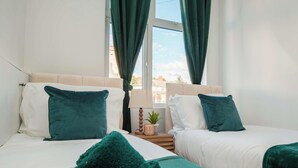 Quarto de hotel com duas camas, cada uma com cabeceira branca e almofadas verde-água. Há uma mesa de cabeceira de madeira com abajur e uma pequena planta em vaso entre as camas.