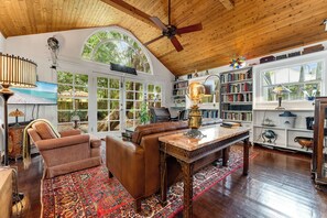 A spacious living room with a high ceiling, wooden beams, a large window, a ceiling fan, a fireplace, a leather sofa, a coffee table, and a bookshelf. - THE WRITER'S COTTAGE ~ Renovated Old Town 2 BR/2 Ba Home w/Parking! (Key West)