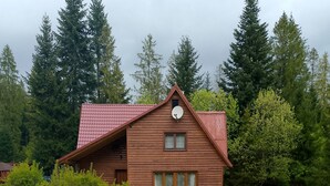 A wooden cabin with a red roof, surrounded by a gravel path, green grass, and trees. - Chata Bobík (Oravice)