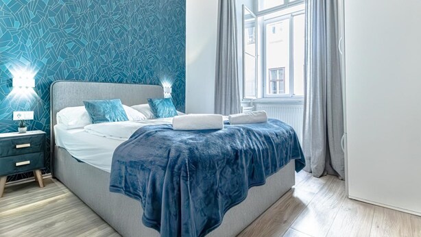 Une chambre à coucher avec un lit, des tables de chevet, une fenêtre avec des rideaux et un mur décoré d’un motif.