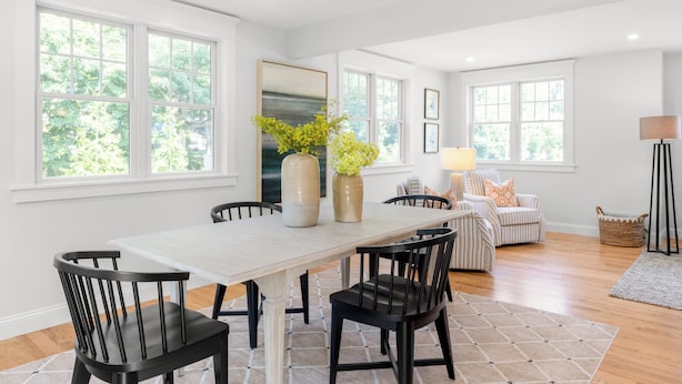 A spacious dining area with a large table, black chairs, and a vase with yellow flowers. There are two windows providing natural light, and a lamp on a side table. - 4-bedroom house in Tisbury for a relaxing getaway - newly renovated (Tisbury)