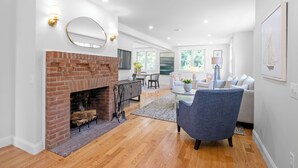 A spacious living room with a brick fireplace, a dining area, and a seating area with a blue armchair and a gray sofa. - 4-bedroom house in Tisbury for a relaxing getaway - newly renovated (Tisbury)