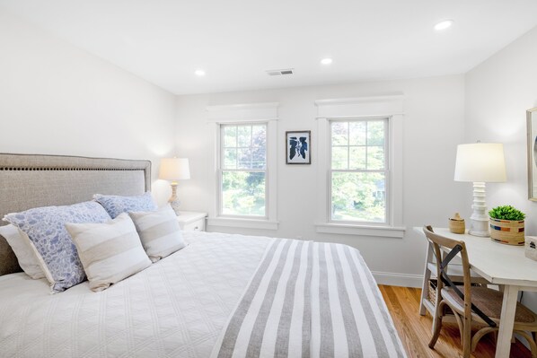 A bedroom with a bed, two windows, a bedside table with a lamp, a chair, and a small table with a plant. - 4-bedroom house in Tisbury for a relaxing getaway - newly renovated (Tisbury)