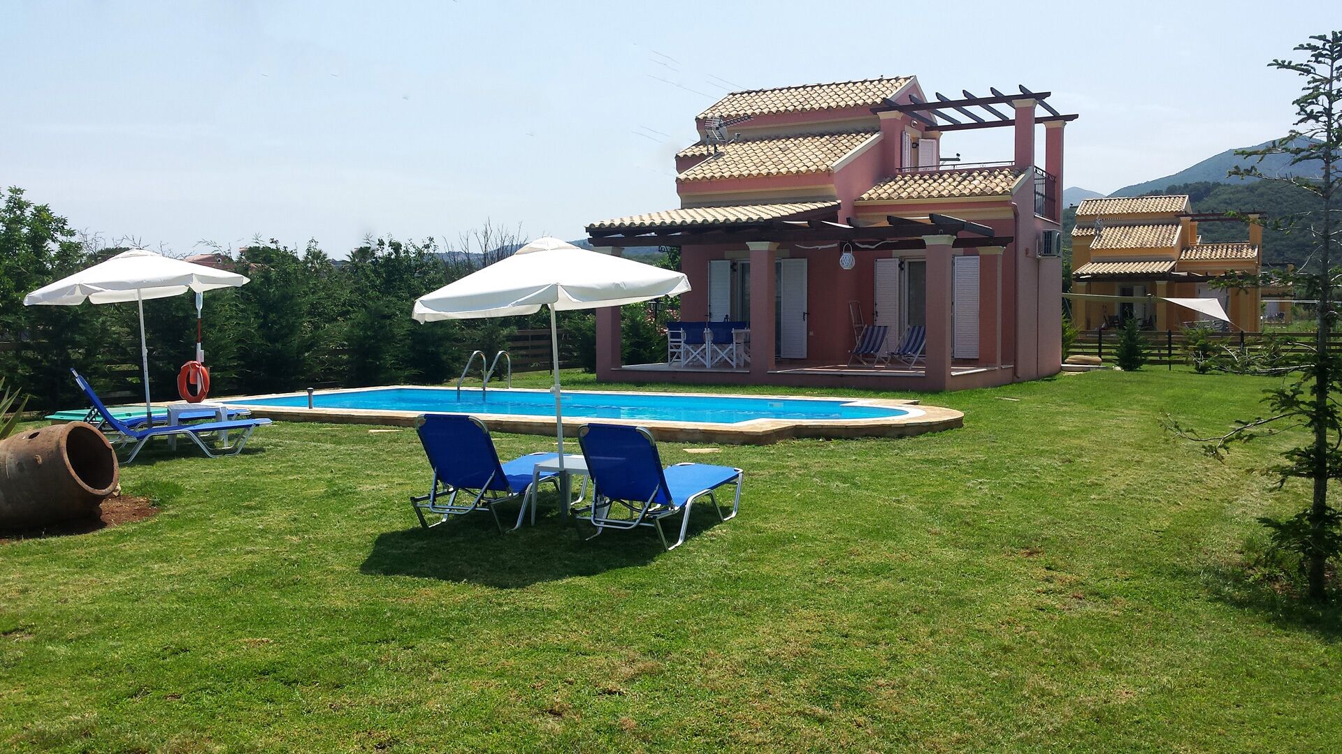 A pool area with blue lounge chairs and white umbrellas, a pergola, and a building with a tiled roof.