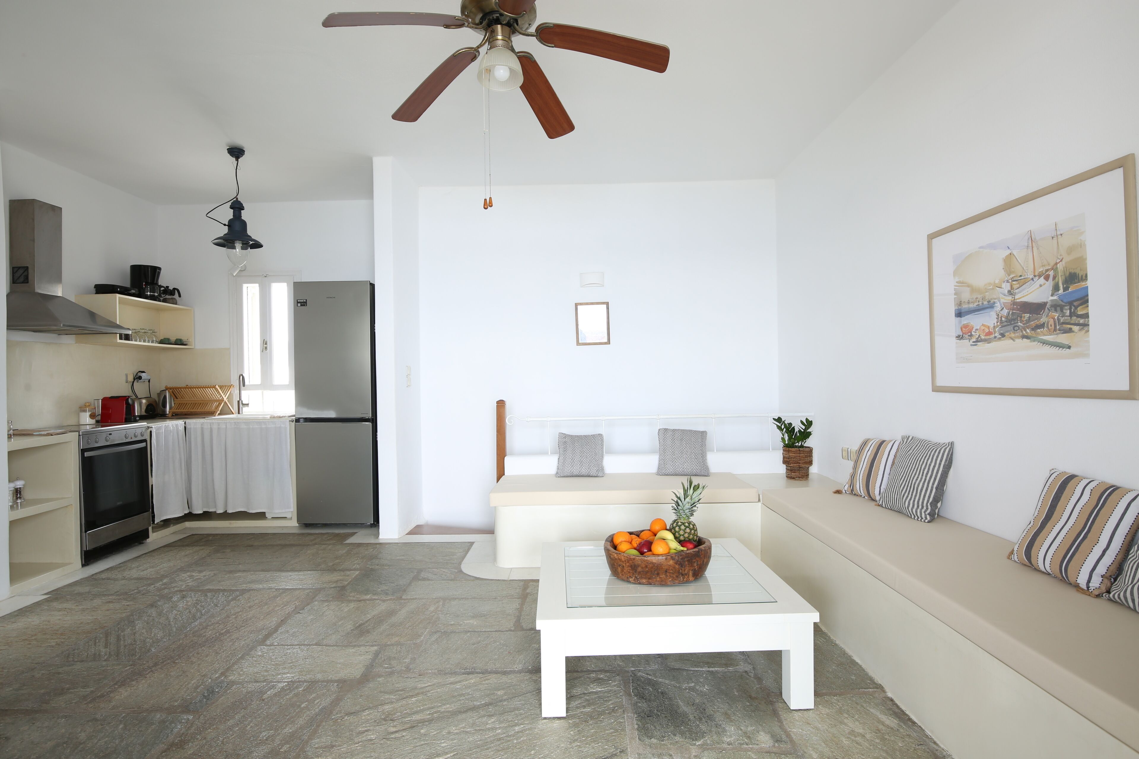A modern kitchen with a built-in oven and refrigerator, a dining area with a white table and bench, and a living room with a sofa and a framed picture on the wall.