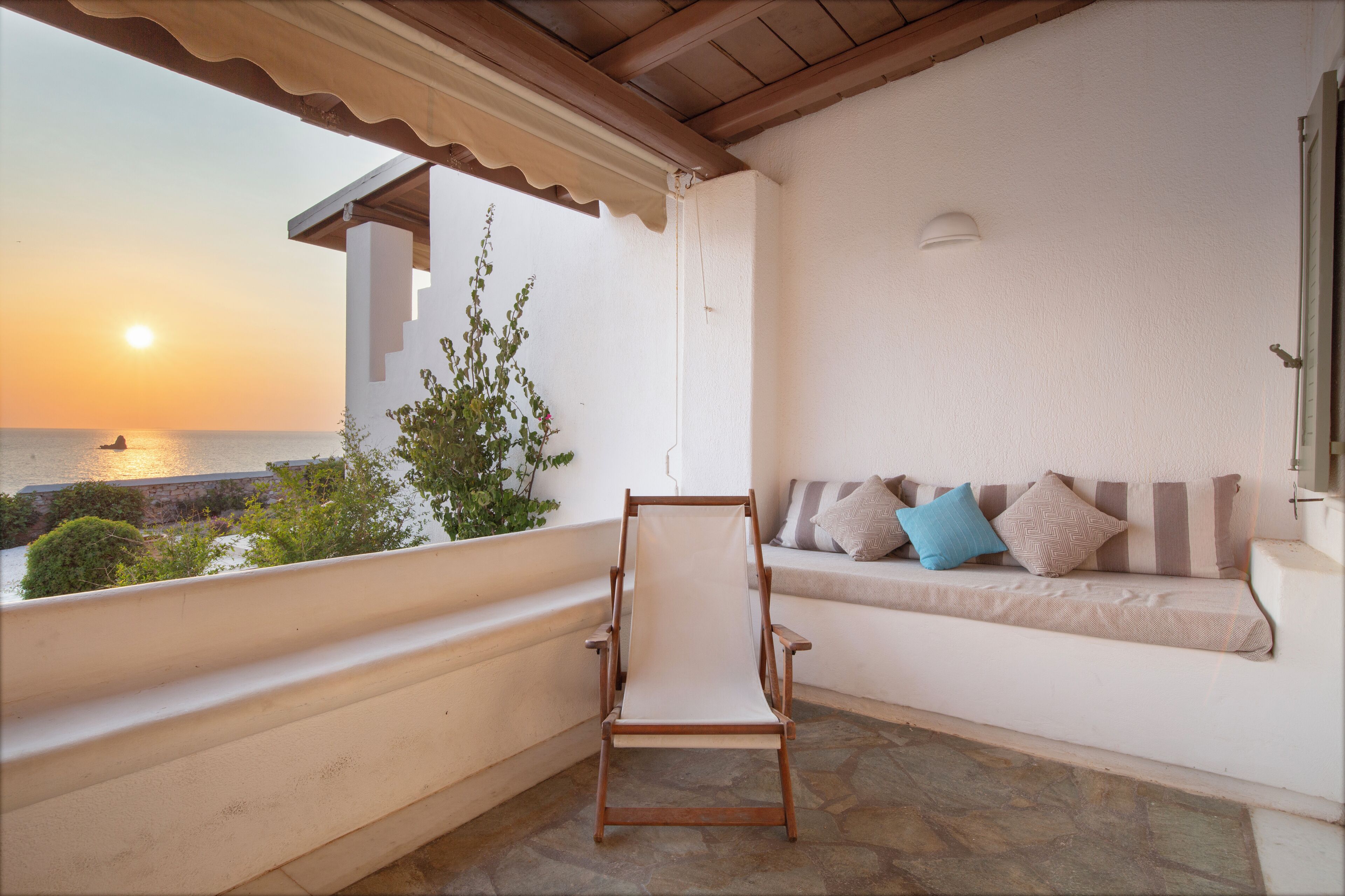 A balcony with a wooden chair, a cushioned bench with pillows, and a view of the sunset over the sea.