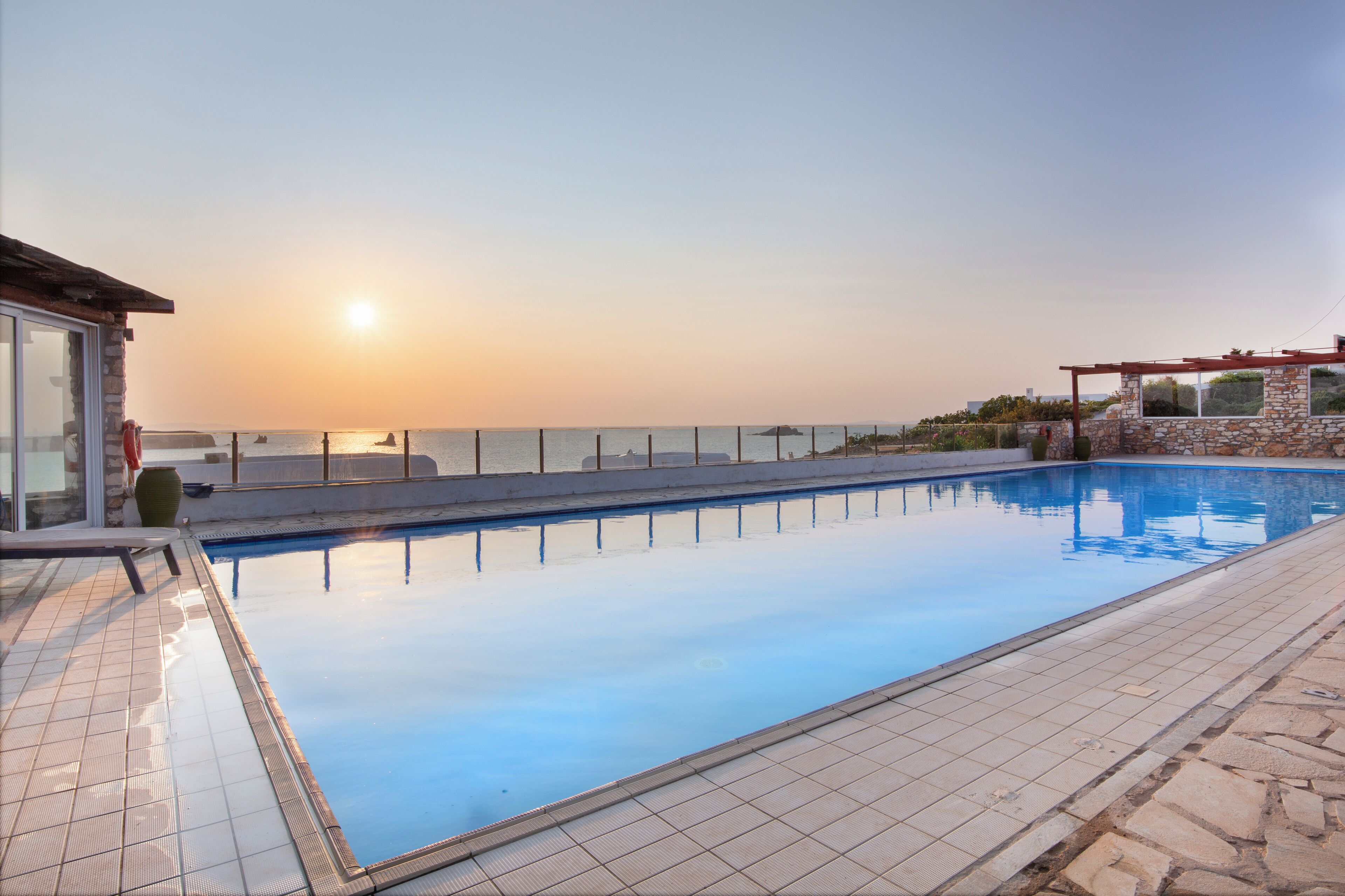 A swimming pool with a view of the sunset over the ocean, a stone wall, and a pergola.