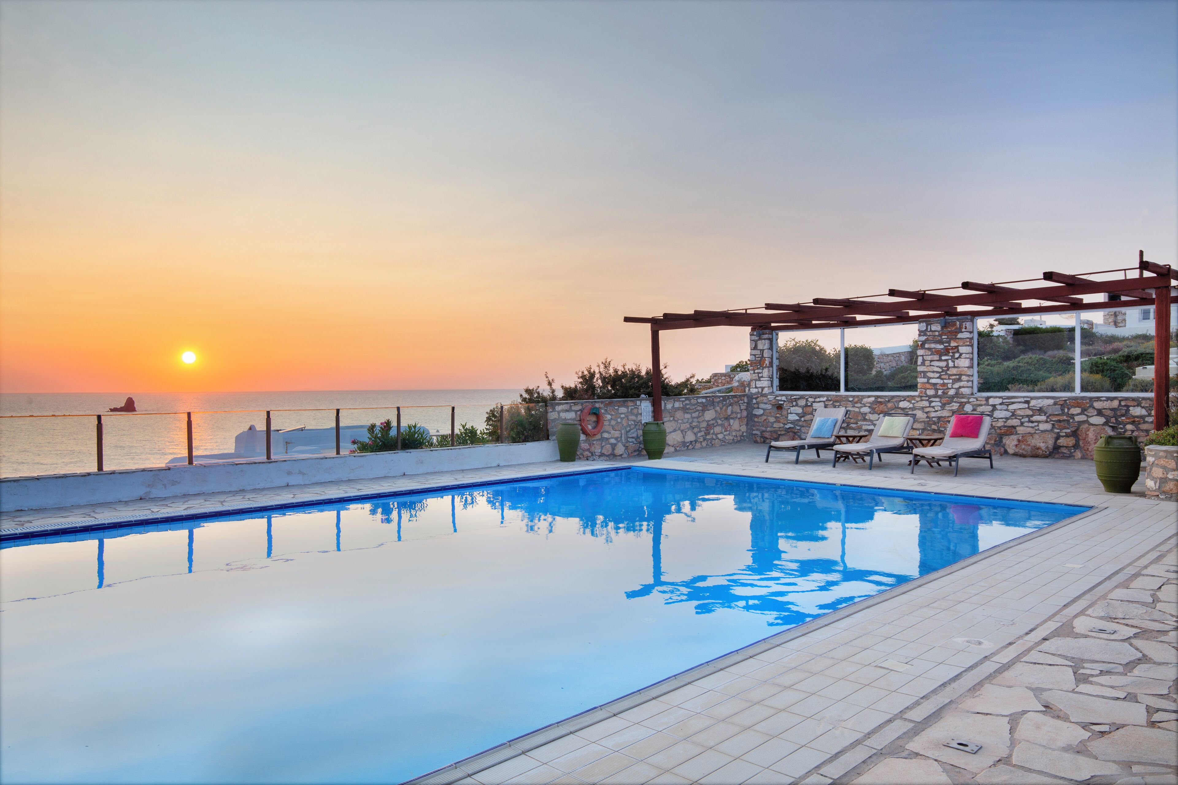 Piscina con pared de piedra y una pérgola con sillas, con vista a un cuerpo de agua al atardecer.