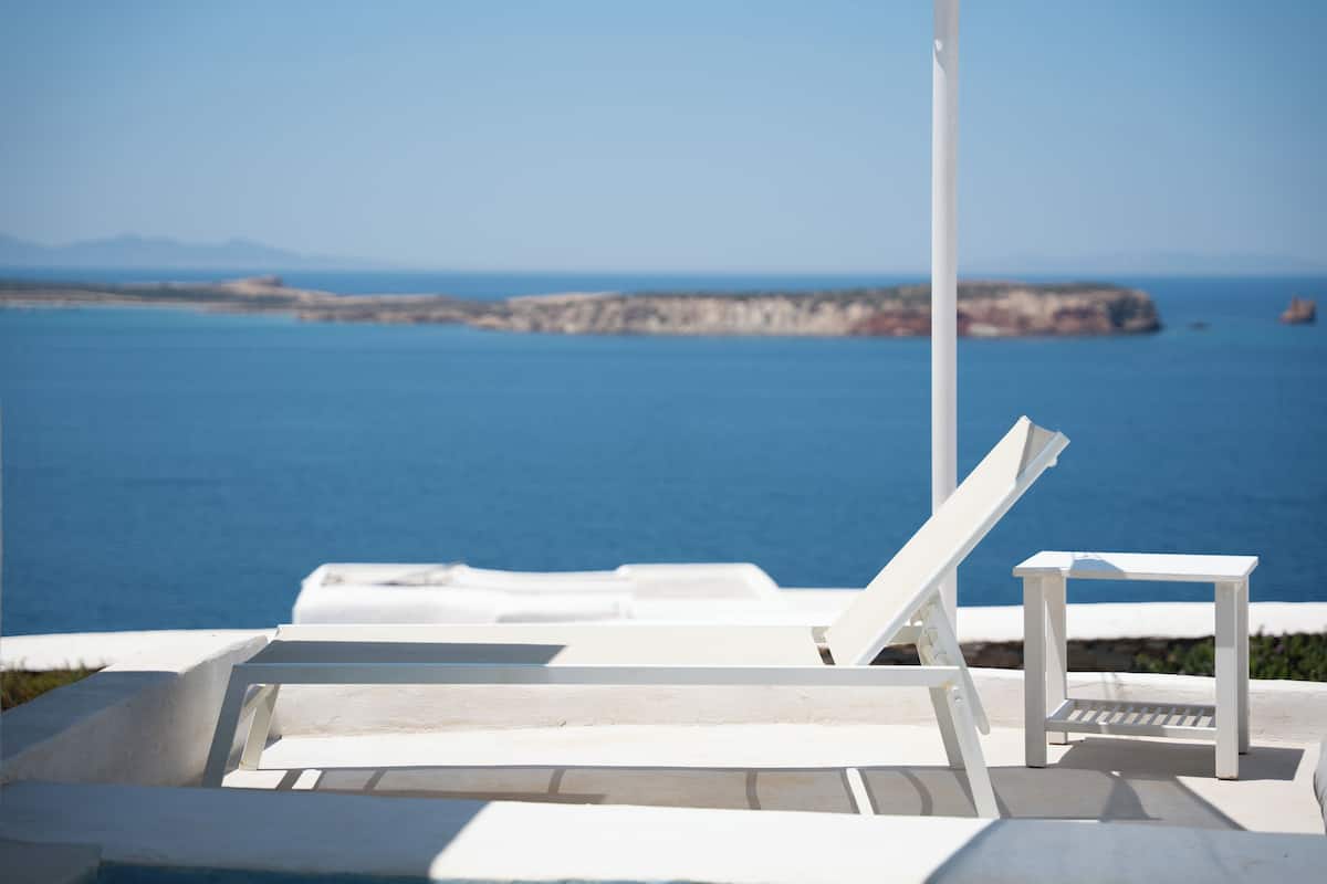 A white outdoor seating area with a view of the sea and a distant rocky coastline.