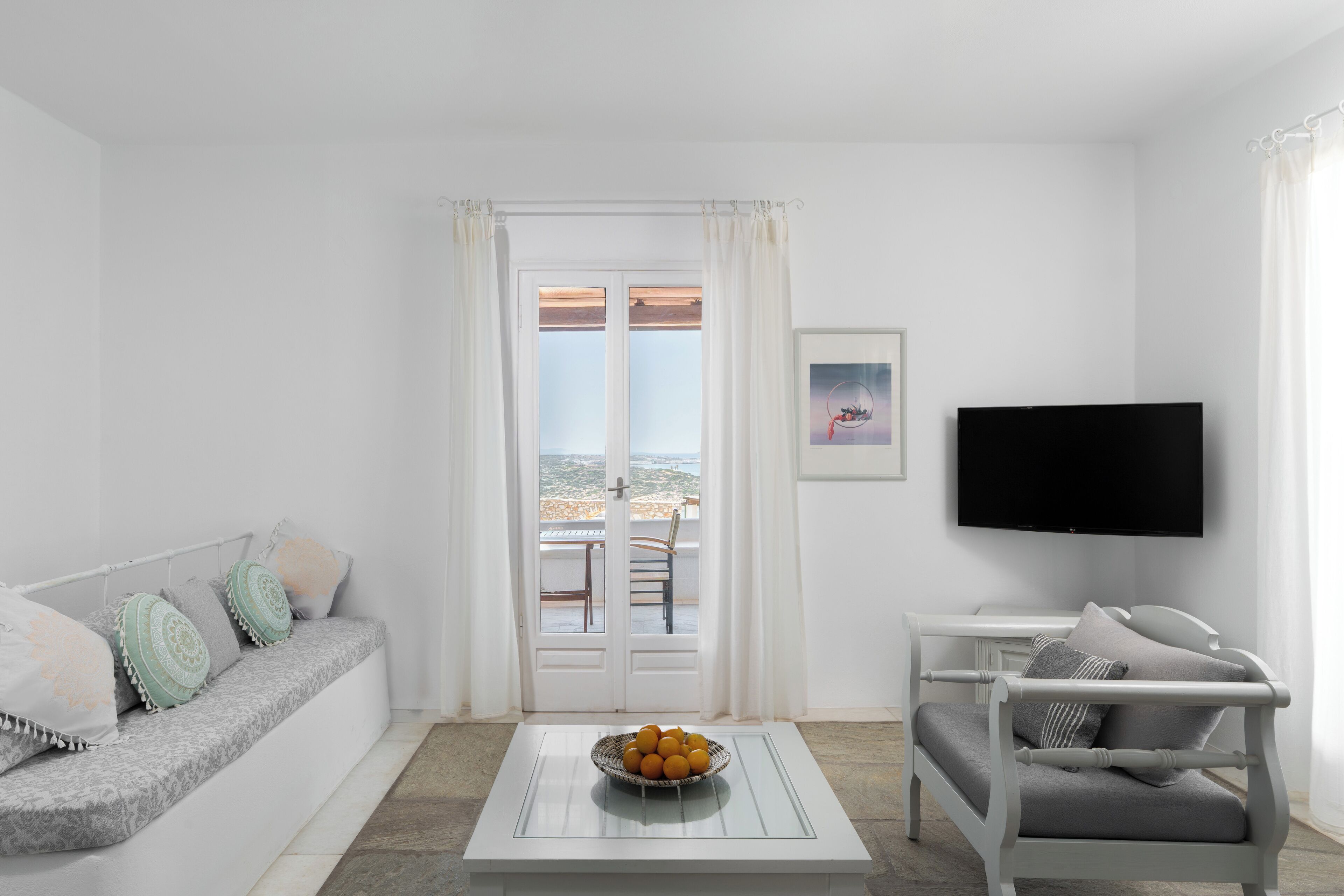 Un salón minimalista con un sofá, una silla, una mesita de centro con un frutero, un televisor y una puerta que da a un balcón.