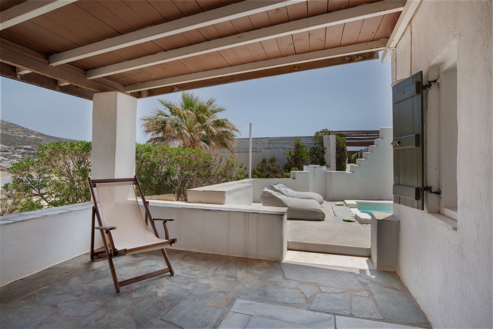 A covered outdoor area with a stone floor, a wooden chair, a small pool, and a view of a mountainous landscape.