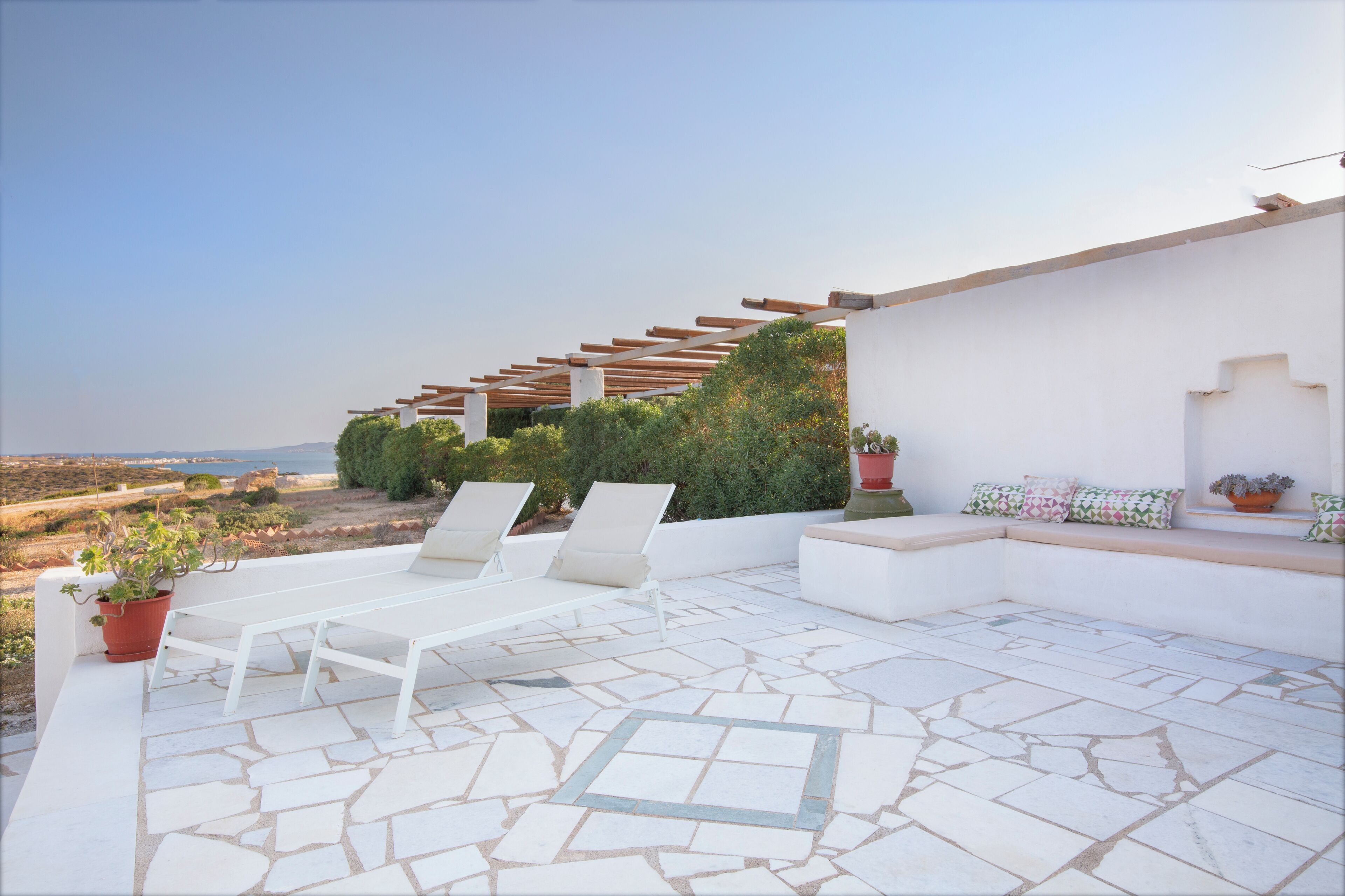 A rooftop patio with white lounge chairs, a pergola, potted plants, and a view of the sea.